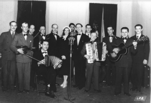 Black and white photograph of the Montagnards Laurentiens in evening dress. Some are wearing bow ties and others are wearing regular ties. A microphone, marked “CHRC,” is in the centre. Lévy Beaulieu is playing the diatonic accordion and Edmond Bélanger is playing the piano accordion.