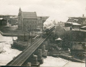 Photograph of a train travelling on a track with Almonte town hall in the background, 1950s