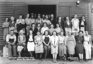 Black and white photo of a group of twenty women and ten men arranged in three rows outside the open door of an old wooden building. Most are looking at the camera. Women are wearing dresses or trousers. The men are wearing trousers or coveralls and collared shirts; some are wearing hats. One man is bare-chested.