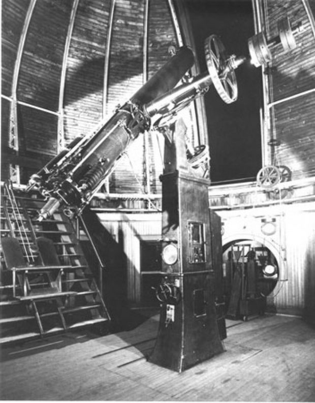 A black and white image of a 15-inch refracting telescope. The telescope is attached to a mounting pier and is inside a domed building. There is an opening in the dome that allows the telescope to view the night sky.