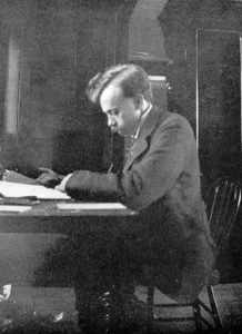 A black and white photograph of a man sitting at a writing desk, He is sitting on a wooden chair and looking at papers on the desk