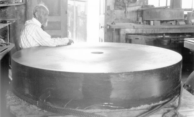 A black and white photograph of a man leaning on a large, thick circular piece of glass