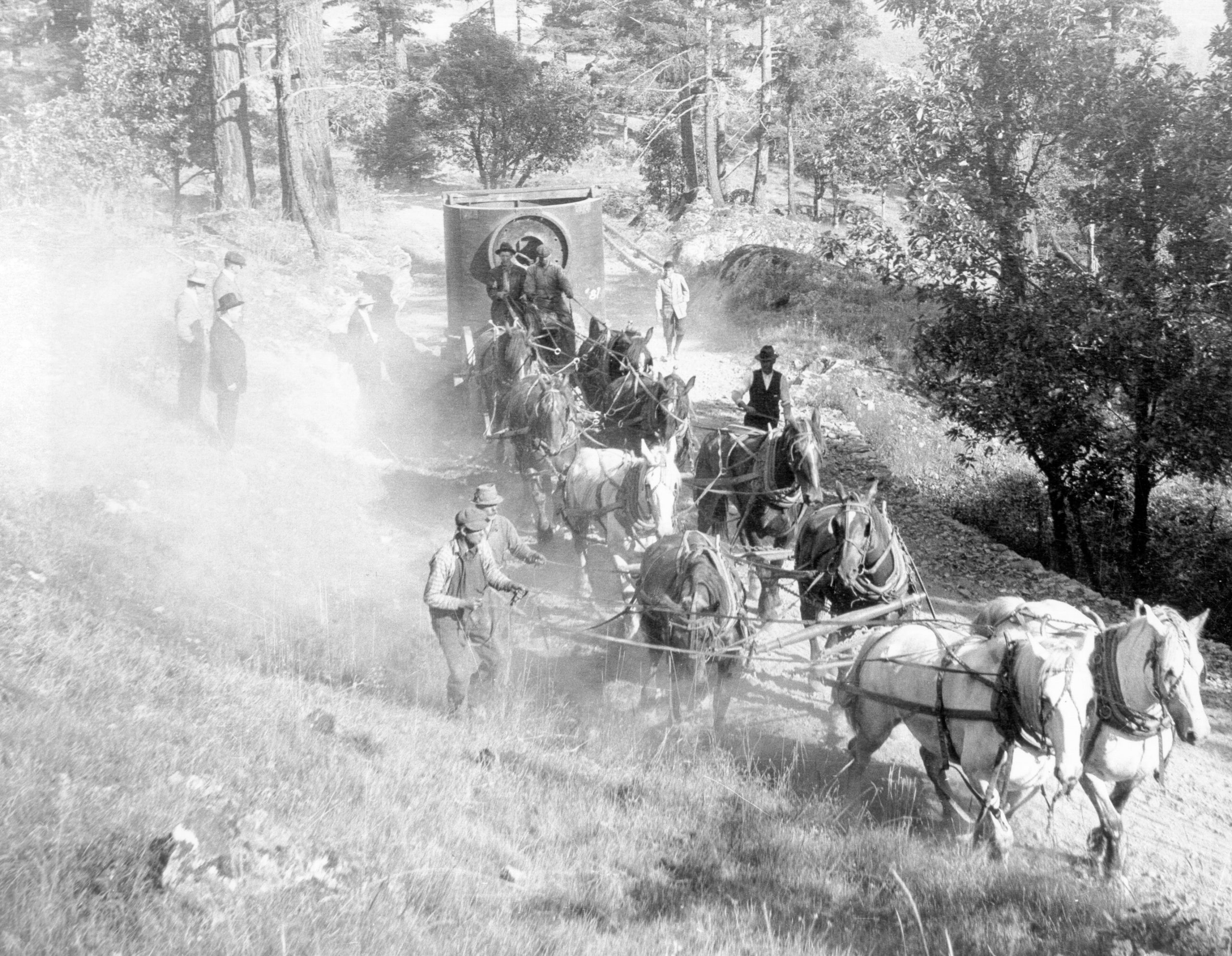 A black and white photograph of a horse-drawn wagon carrying a very large piece of equipment up a dusty hillside road