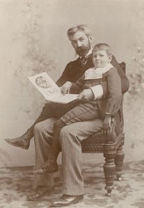 A sepia colored photograph of a man holding a book sitting on a chair with a young boy sitting on his lap