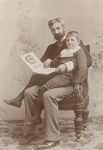 A sepia colored photograph of a man holding a book sitting on a chair with a young boy sitting on his lap