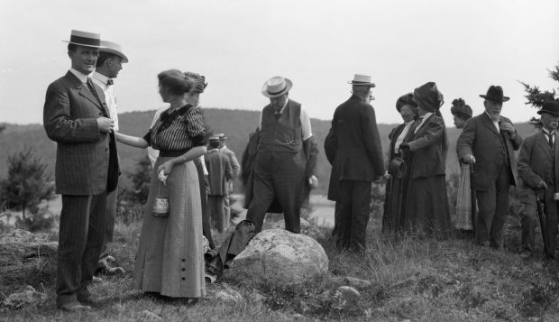 A black and white 1911 photograph of a group of people on a hilltop standing and talking