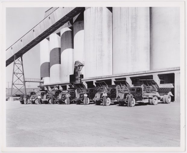 A row of rock hauler trucks and their drivers set against a quarry building