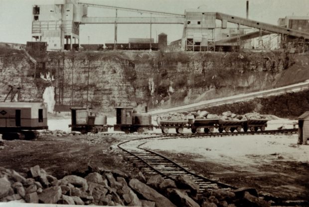 A small train and four carts are seen along a track inside a quarry pit. In the background are several industrial structures.