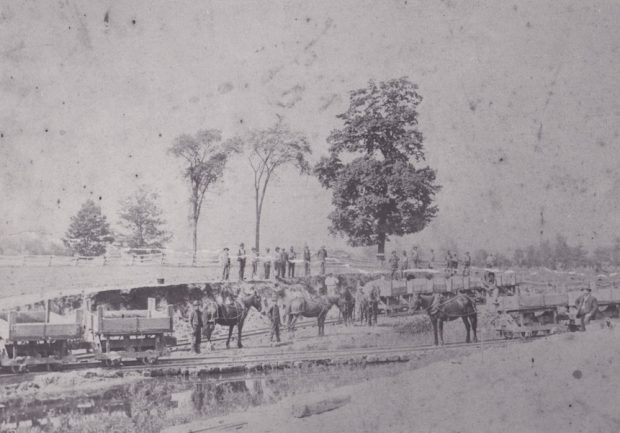 A faded black and white photograph of a work crew outside looking down on a group of horses attached to wooden carts along a track