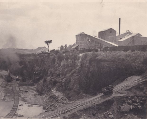 A cart makes its way up a track on a ramp that leads out of a pit to a set of industrial buidlings.