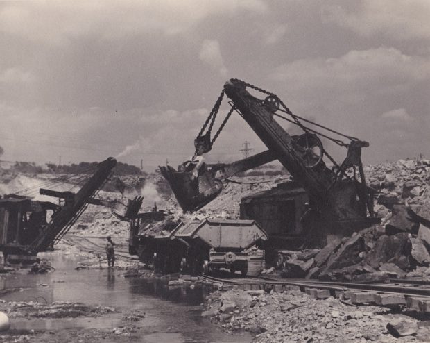 A big piece of machinery hangs its bucket over carts waiting along a track.