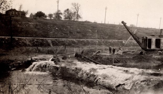 An image of two people standing beside a steam shovel and a pump pouring water into a pond