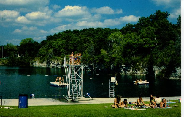 People at quarry lake: swimming, waiting to jump off a high dive, and lounging on the grass.