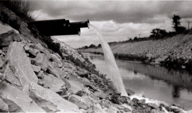 Several black pipes extend out of a heap of stones and direct water to a river below them