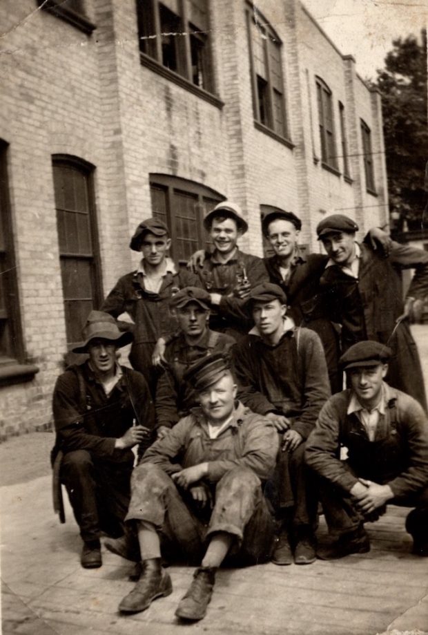 A group of 9 young men huddle together in their work clothes and hats