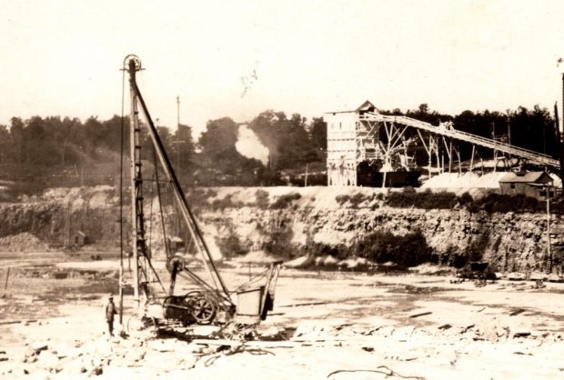 Black and white photograph of a man standing in a pit in the earth beside a towering drilling device