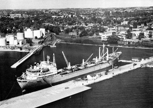 a black and white photo of an industrial harbrourfront with a large tanker vessel at dock in the middle, white oil tanks to the left, railway lines and piles of coal in the middle and a boxcar to the right. In the right distance is Victoria Hall.