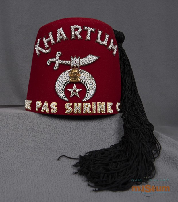 A red Shriners fez. On top there are four sets of air holes in the form of circles, and three rows of holes in each circle. The beaded lettering on the front reads KHARTUM THE PAS SHRINE CLUB. There is also a symbol of an Arabic Sword and an Egyptian crest with a silver star below it. A long black tassel is attached from the top and hangs down the side. On the right side there is a silver broach with a studded Arabic sword and a crescent below with a maple leaf in the center.