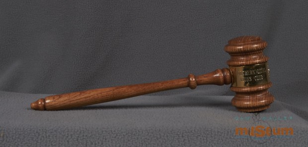 This gavel has a handle attached to a mallet style head, both made of wood and lightly stained with a varnish layer. Both parts are carved with simple lathe style designs. The middle of the mallet head is wrapped with a sheet of metal and the ends are fixed where the handle attaches to the mallet head. The metal sheet has the Lions emblem and the text: Northern Lites/Lions Club and The Pas/Manitoba.