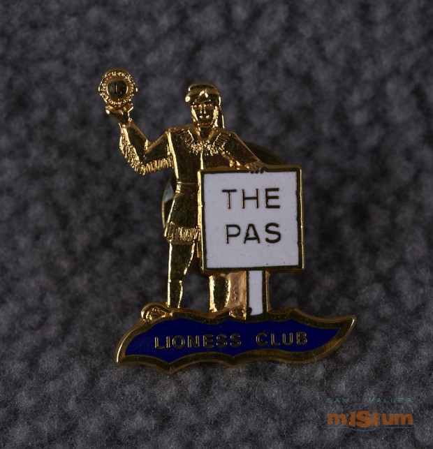 This pin is in the shape of the Trappers sign. The recto shows the man from the Trappers sign, in gold, holding the Lions emblem in his hand. There is a white sign in front of him with gold coloured lettering reading The Pas. He and the sign are standing on an irregular shaped piece with a dark blue field.