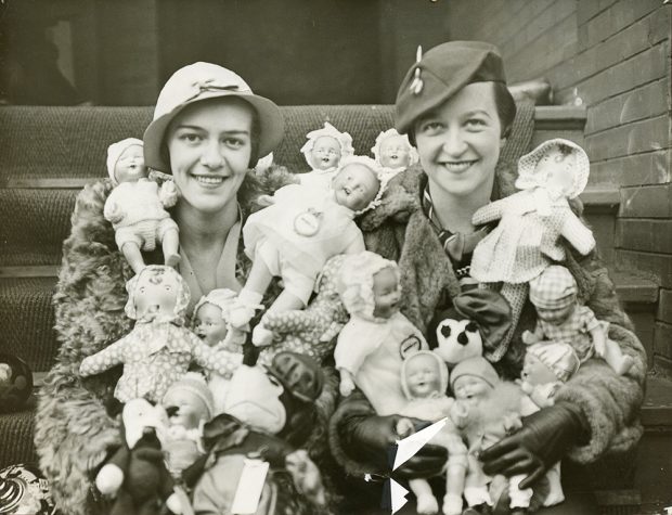 Two fashionably dressed women with arms full of baby dolls sitting on outdoor steps in a black and white photo.
