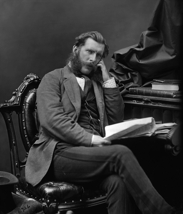 Black and white photographic portrait of Dr. John Sweetland at about age 35, sitting on a leather chair in front of a desk. He is wearing a suit and holds a book open.