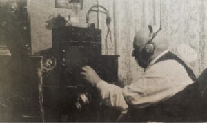 Black and white photograph of a man with headphones sitting in a chair beside a radio. He is wearing a white shirt and a black vest.