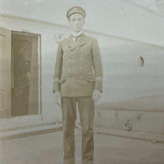 Sepia photograph of a young man in a uniform standing on the deck of a ship.