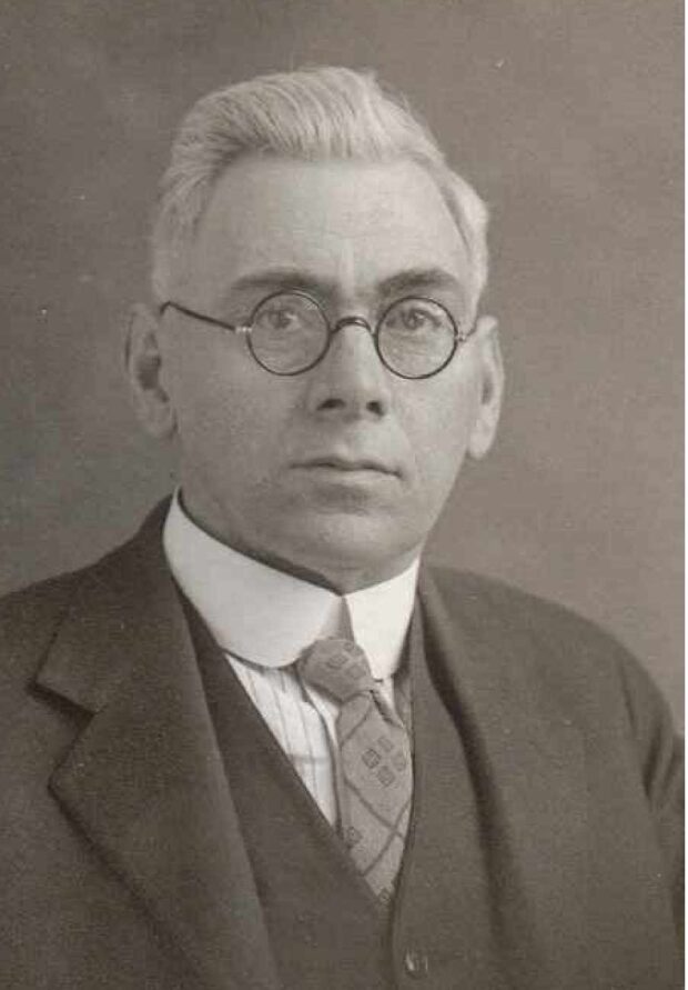 Black and white photographic portrait of a man with grey hair and glasses. He is wearing a white collared shirt, tie, and blazer.