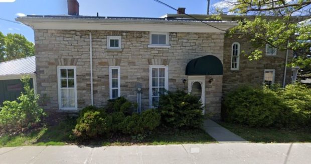 Colour photograph of the front view of the Doctor's House from May 2023. It is a stone house with white doors and windows, and shrubbery in front of the windows.