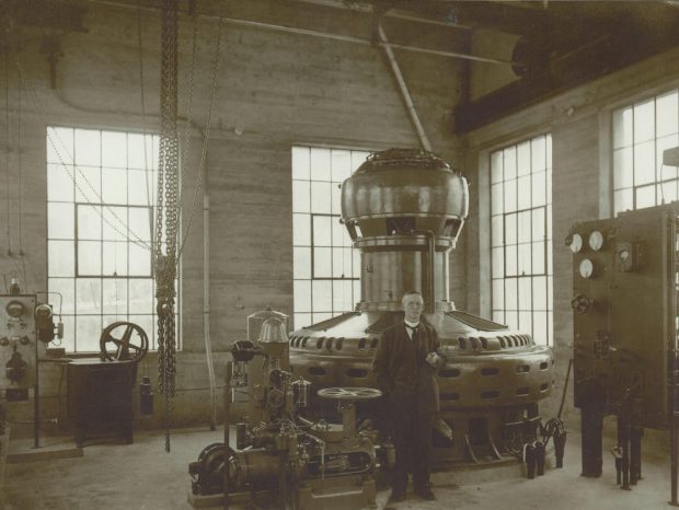 Black and white photograph of a man in a suit standing in a large room with machinery.