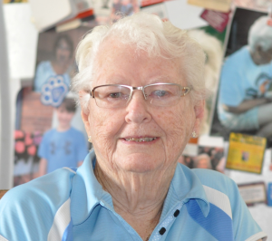 Colour photographic portrait of a woman with white hair who is wearing glasses and a blue shirt.