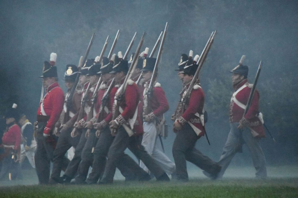 A photograph of re-enactor soldiers in 1812 uniforms marching in the rain.