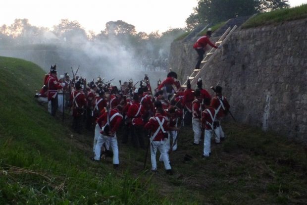 A coloured photograph of re-enactor soldiers in a ditch before a stone wall scrambling up a wooden ladder.