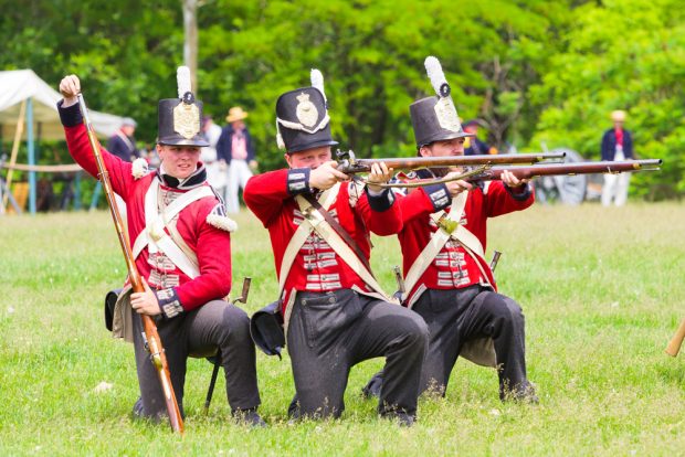 A coloured photograph of three kneeling 1812 re-enactor soldiers, two of which are firing and one reloading.