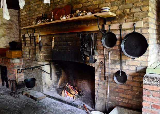 A coloured photograph of a stone fireplace with a small, glowing wood fire. Three iron frying pans hang to the right of the hearth. A simple shelf above the hearth contains jugs and cups.