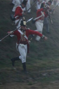 A soldier, carrying a pike and with other soldiers, charges down a slope in the dark, shouting.