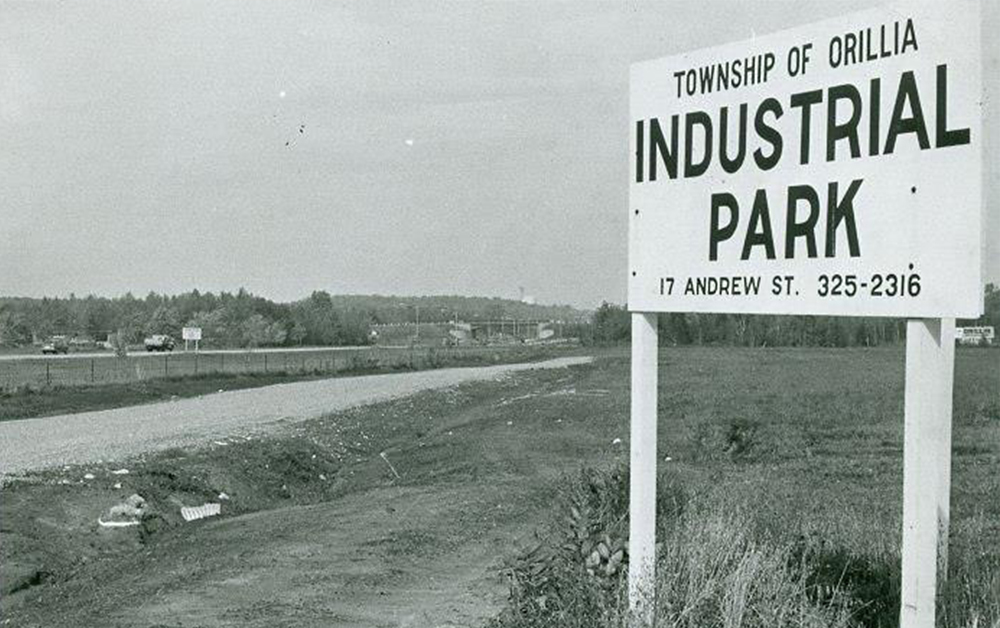 Vintage black and white photograph circa 1968. Large, white, wooden road sign in the countryside reads, 