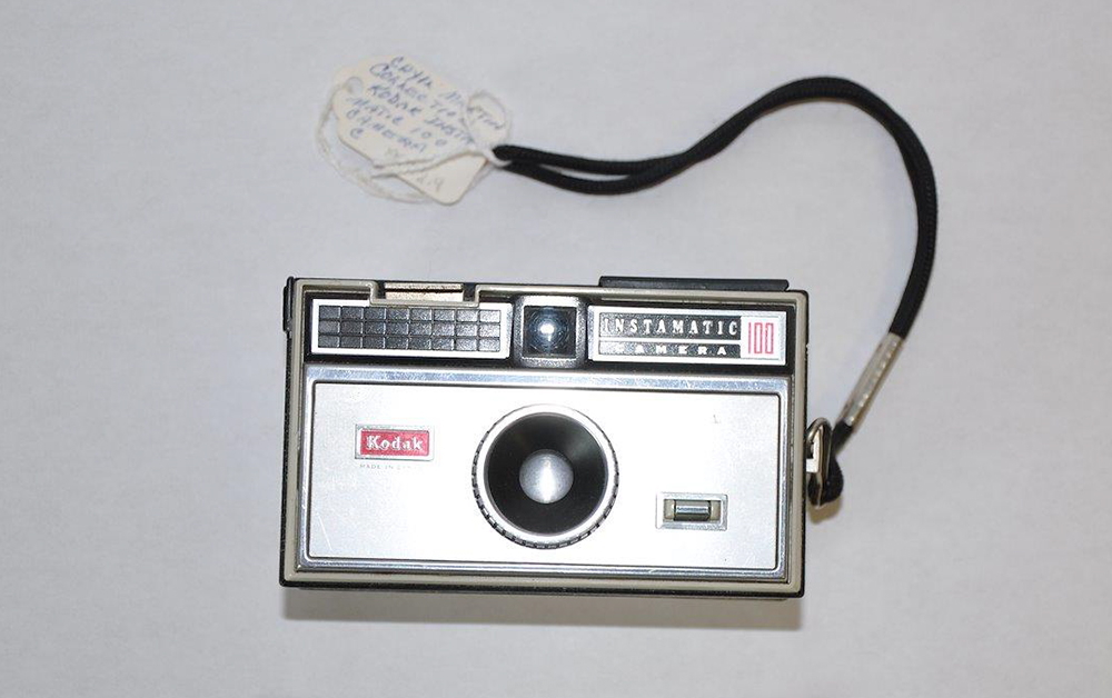 Small, rectangular, silver Kodak Instamatic Camera 100 with red details and black wrist strap, circa 1963.