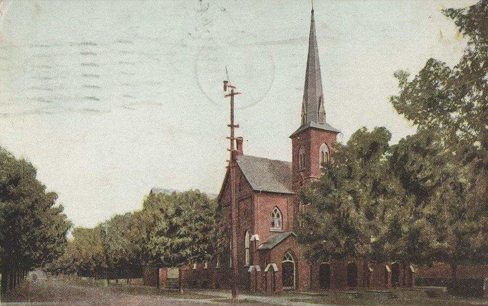 Colour-tinted postcard of a rural Orillia steeple church built with red brick, along a densely tree-lined street, circa 1909.