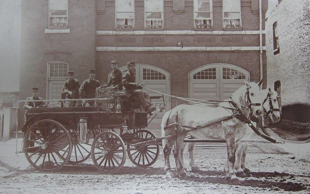 Vintage sepia photograph circa 1900. Orillia Fire Brigade in front of brick Fire Hall. Five men sit atop an industrious fire carriage, lead by two white horses over a sandy road.