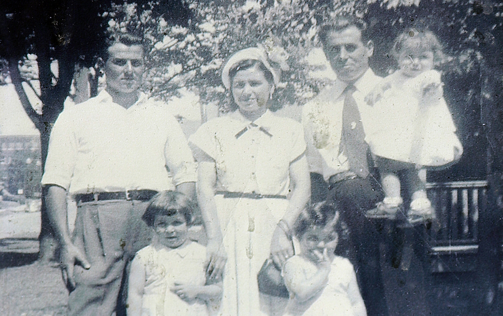 Vintage black and white photo circa 1953. Two men, a woman, and three small girls in white dresses, pose closely together on a tree-lined sidewalk.