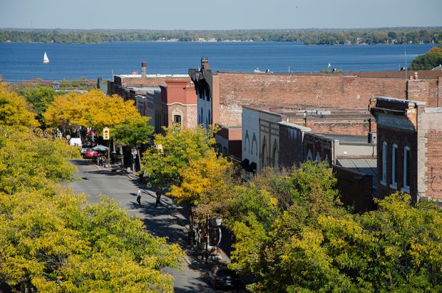 Scenic aerial view of downtown Orillia, overlooks historic red brick buildings, running along a tree-lined street, towards a striking blue Lake Couchiching.