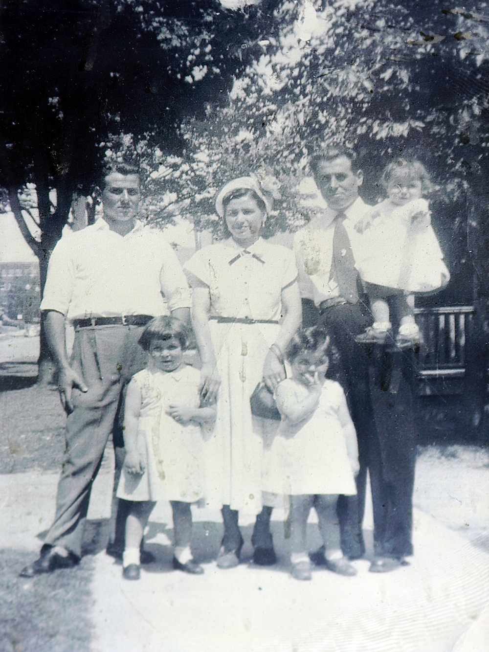 Vintage black and white photo circa 1953. Two men, a woman, and three small girls in white dresses, pose closely together on a tree-lined sidewalk.