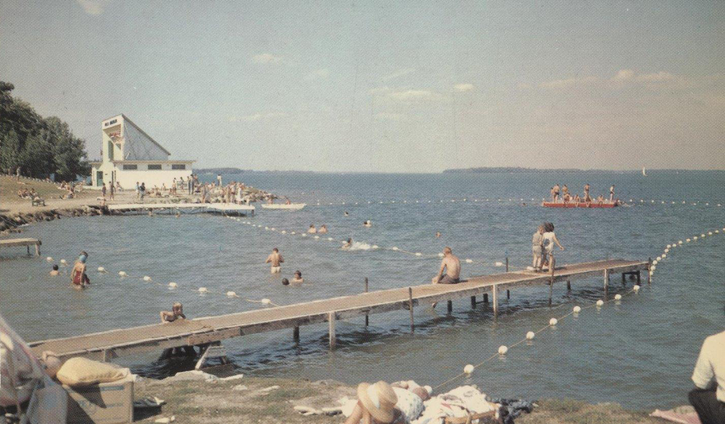 A vintage colour postcard circa 1960 at Lake Couchiching beach. The water's edge is filled with people swimming, sitting on docks, sunning on the sand, with Orillia's iconic Aqua Theatre back left.