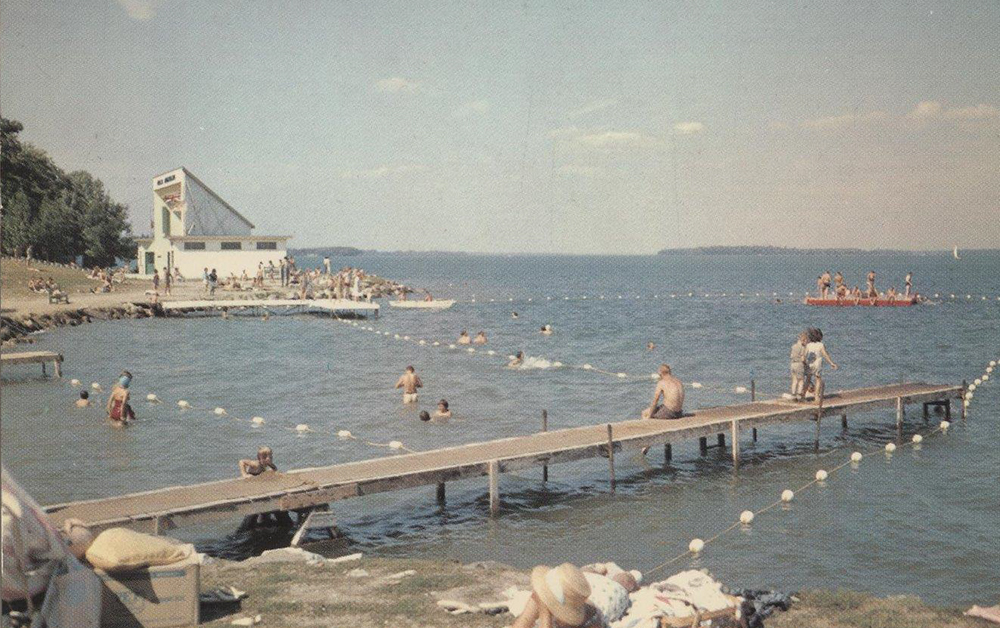 A vintage colour postcard circa 1960 at Lake Couchiching beach. The water’s edge is filled with people swimming, sitting on docks, sunning on the sand, with Orillia's iconic Aqua Theatre back left.