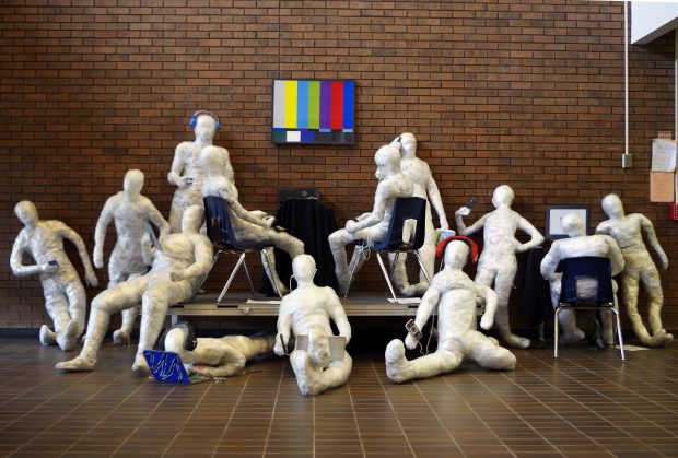 Ten white, human-sized sculptures are strategically placed in front of a red brick wall connecting with laptops and cellphones instead of each other.