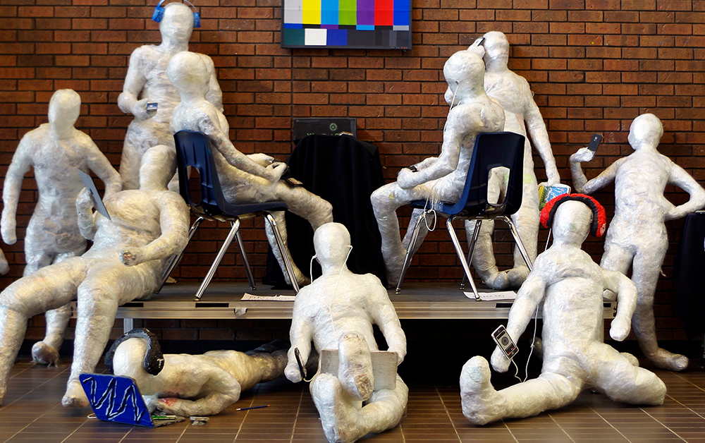 Ten white, human-sized sculptures are strategically placed in front of a red brick wall connecting with laptops and cellphones instead of each other.