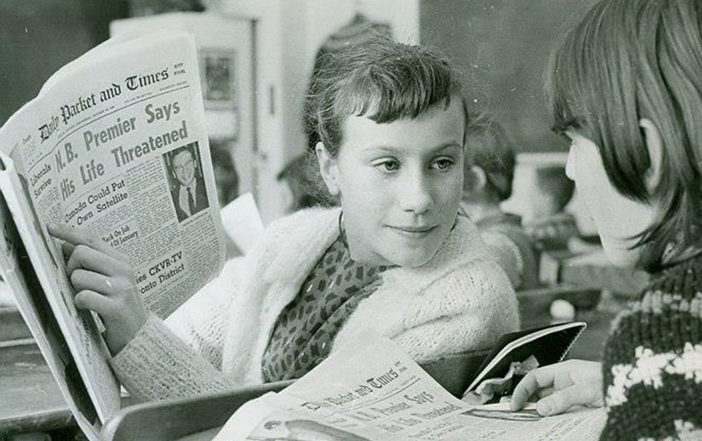 Vintage black and white photograph circa 1960 of two young school girls reading The Packet and Times together in class. One girl points to an article as the other takes notice.