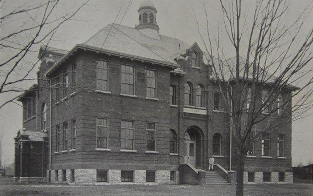 Aged black and white photograph of a man dressed in white sitting at the entrance of a large brick building. Written below the image is 